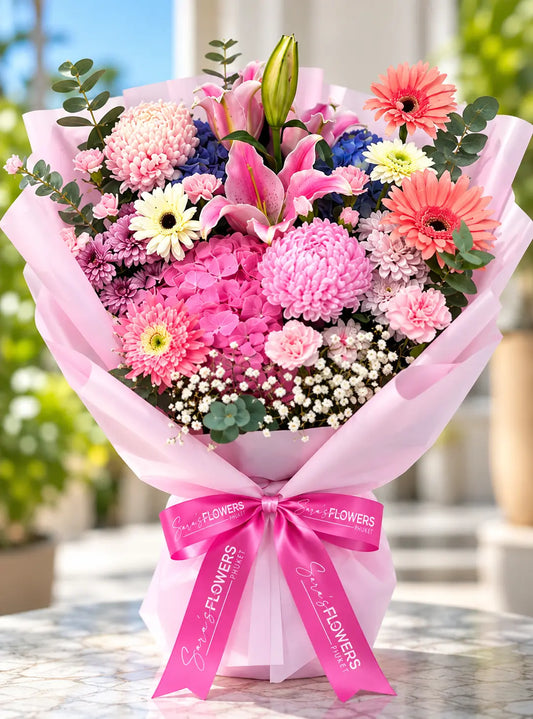 A vibrant flower bouquet wrapped in light pink tissue paper and tied with a bright pink ribbon printed with 'Flowers Direct', set against a softly blurred outdoor garden background.