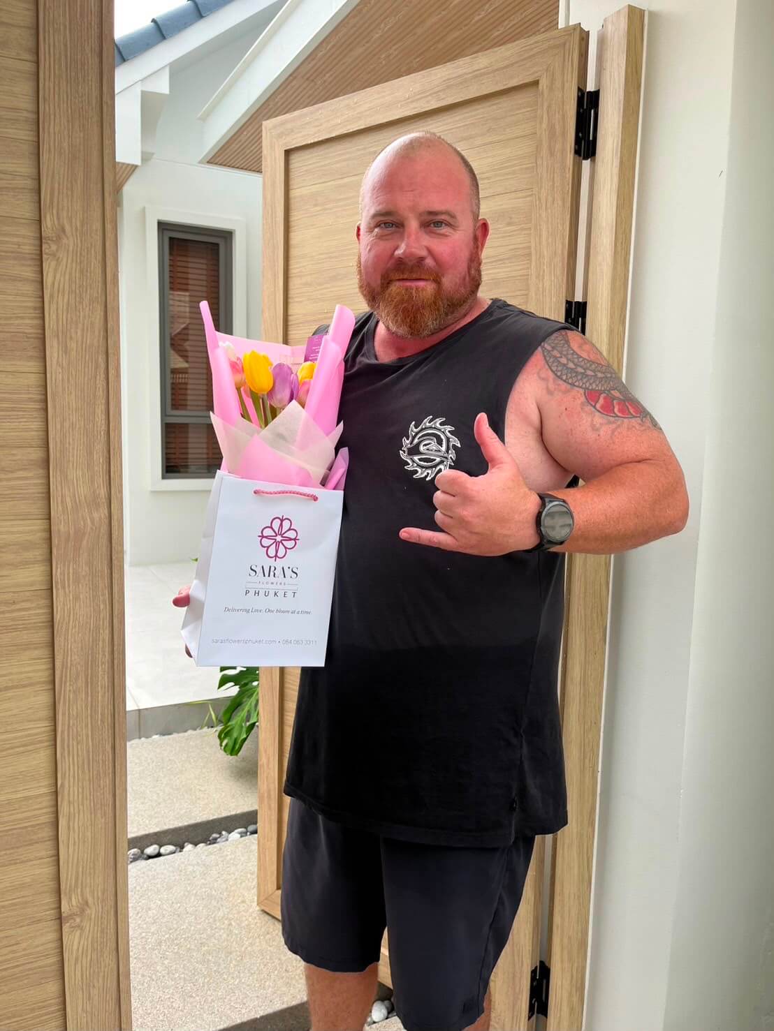 Man holding a bouquet of flowers and a gift bag, standing in front of a door.