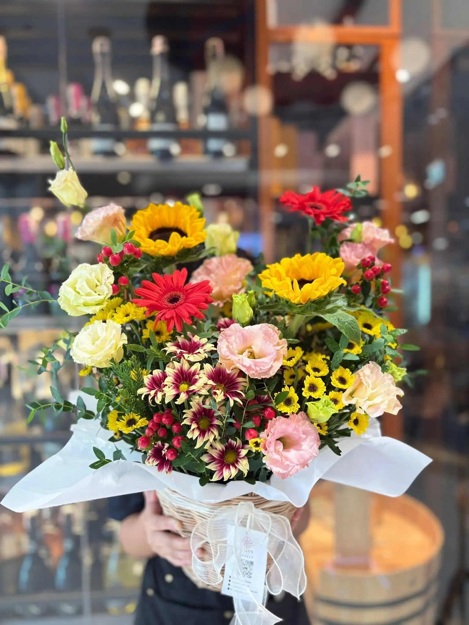 A vibrant multicolor flower bouquet held by a person in front of a softly blurred shop window. The bouquet features yellow sunflowers, red and pink roses, marigolds, and burgundy daisies wrapped in white tissue paper with a sheer white ribbon.