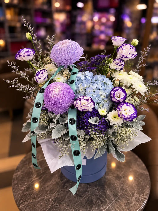 A floral arrangement in a round blue box on a marble table, featuring purple and lavender flowers, white and light blue hydrangeas, green foliage, and a mint green ribbon with black polka dots.