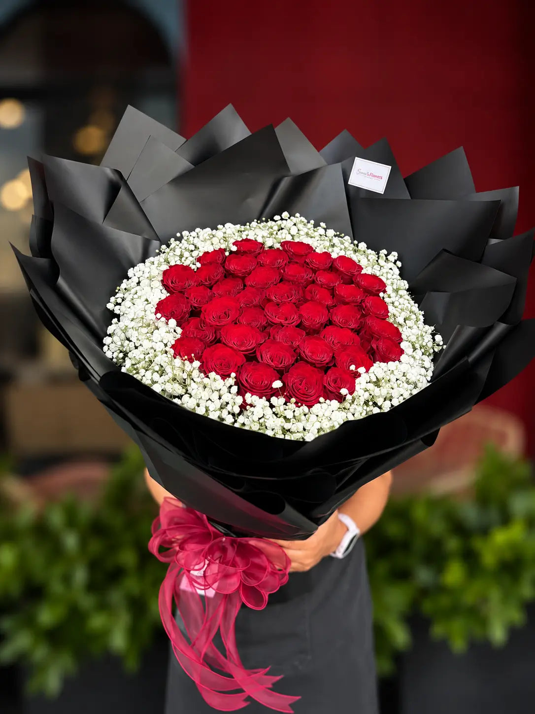 A large bouquet of 39 red roses  surrounded by white baby's breath, wrapped in black paper with a pink ribbon, held by a person wearing a dark apron.