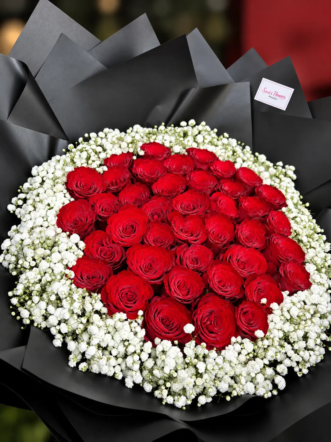 A bouquet of red roses surrounded by white baby's breath, wrapped in black paper with a small card attached.