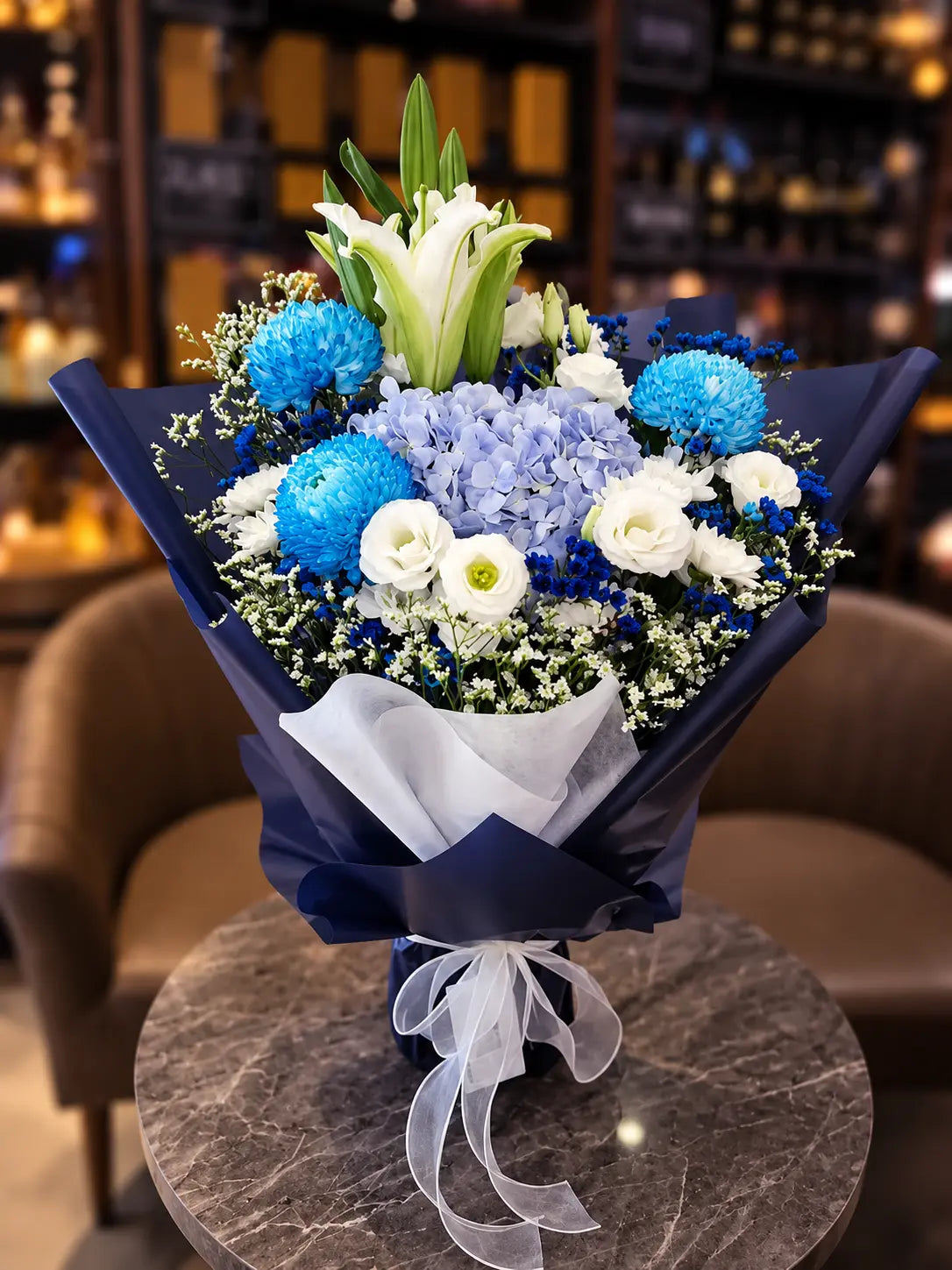 A beautifully arranged floral bouquet featuring blue hydrangeas, white roses, blue pom-pom chrysanthemums, and white lilies, wrapped in dark navy blue paper with a white organza ribbon, displayed on a marble table in a warm, softly blurred indoor setting.