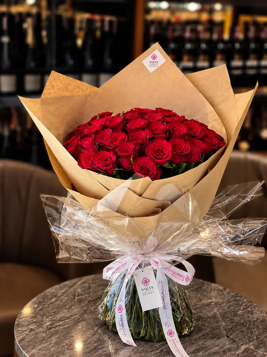 A bouquet of 50 red roses wrapped in brown kraft paper and clear cellophane, tied with a white ribbon printed with the brand name 'Amicus Florist', set on a marble table with a blurred wine shop background.