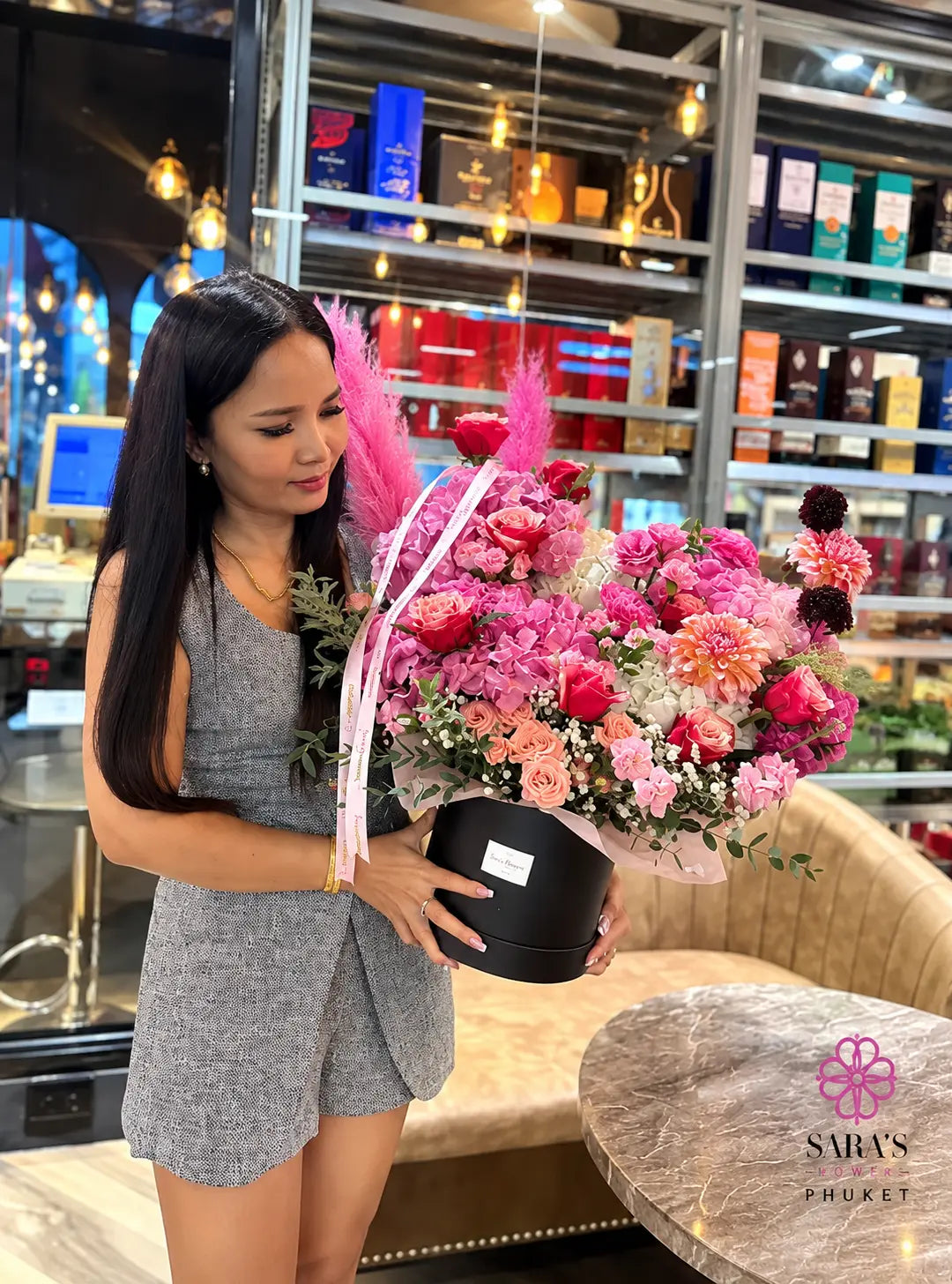 A woman in a gray romper holds a large, vibrant pink and red floral arrangement in a black round box inside a shop. The bouquet features pink carnations, roses, peonies, and pink feather plumes tied with a pink ribbon.