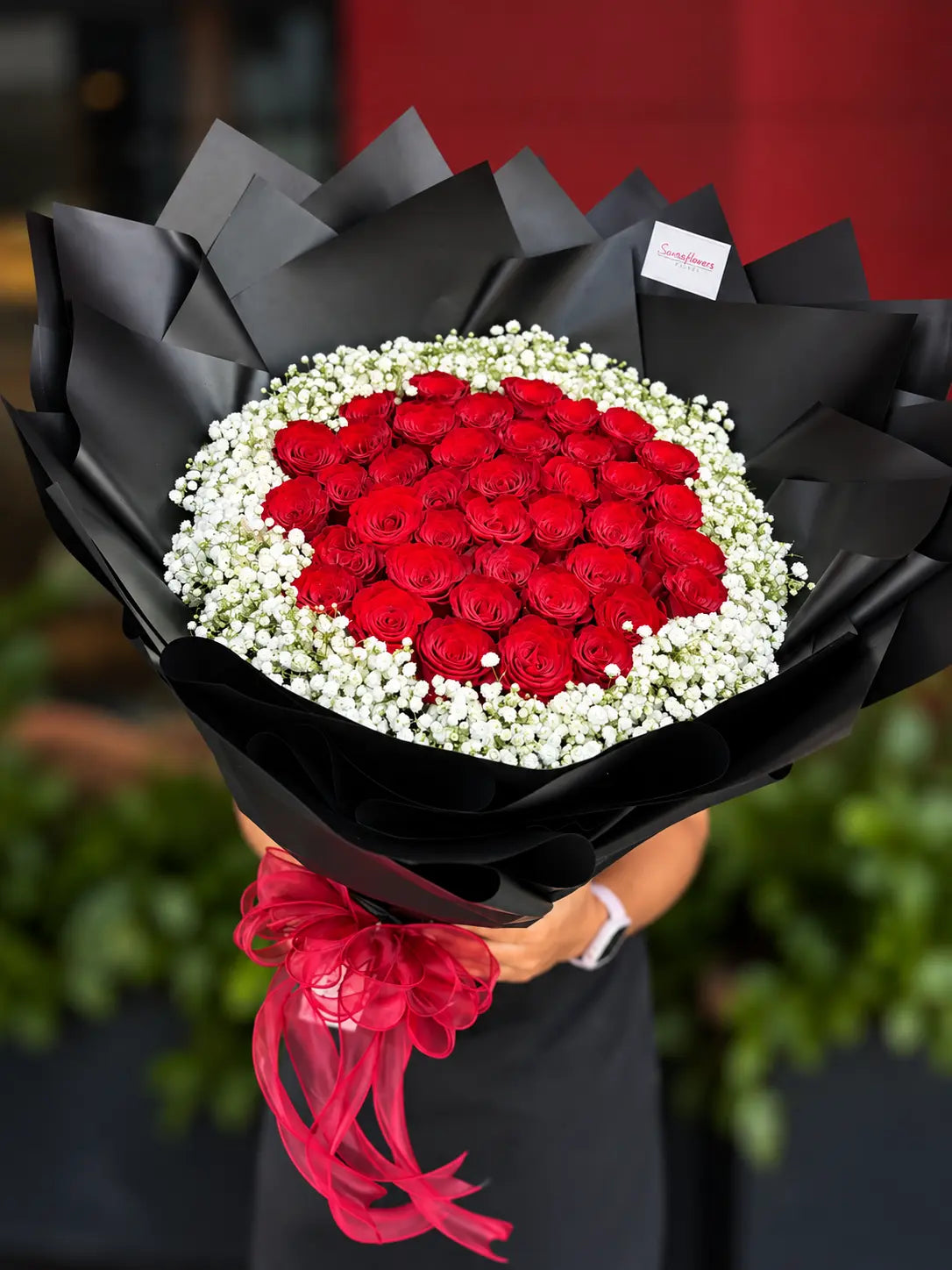 A large bouquet of 39 red roses arranged and surrounded by white baby's breath, wrapped in black paper with a red ribbon, held by a person.