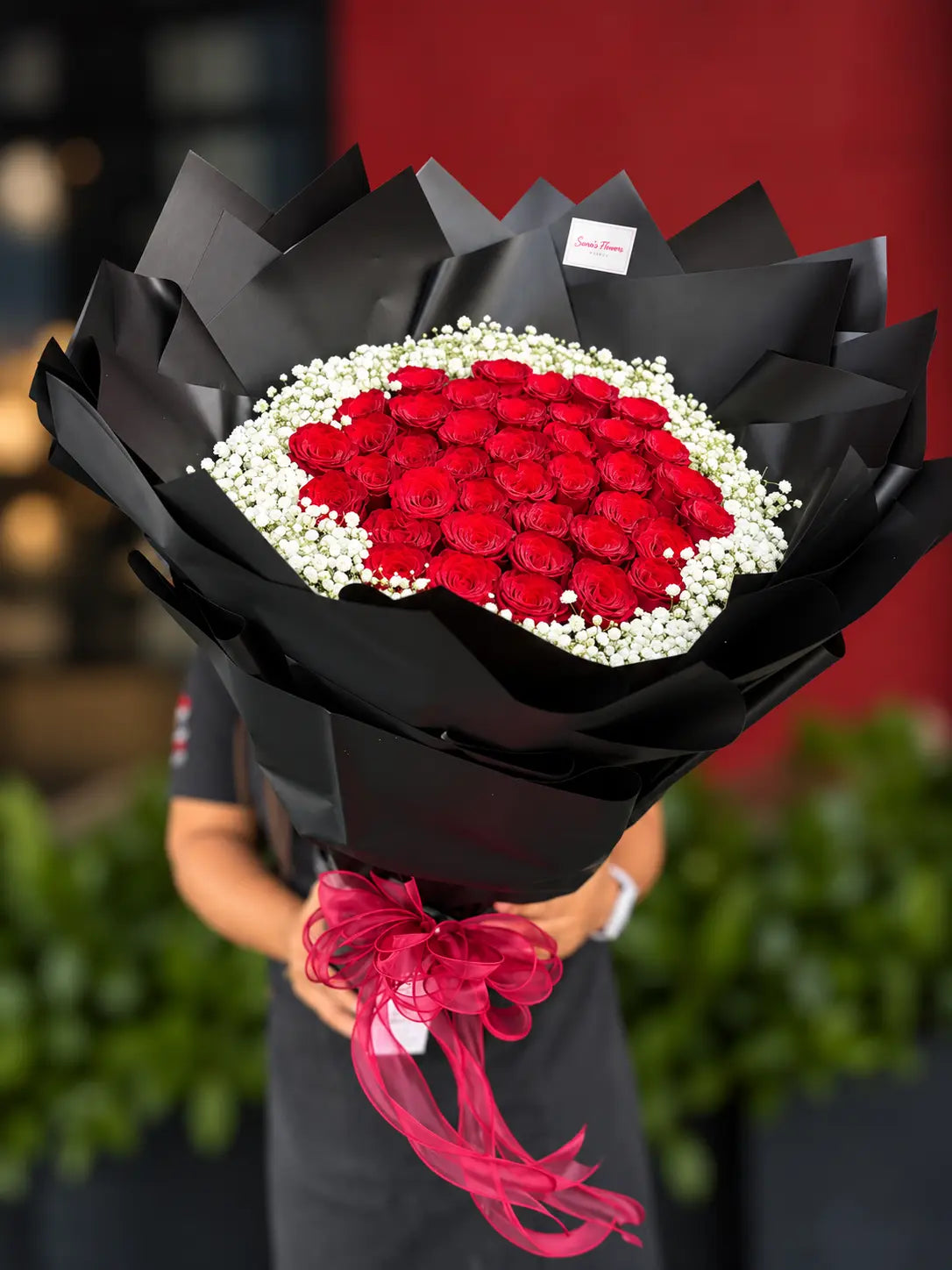 A large bouquet of red roses wrapped in black paper with a bright pink ribbon, held by a person in a dark uniform against a blurred red and green background.