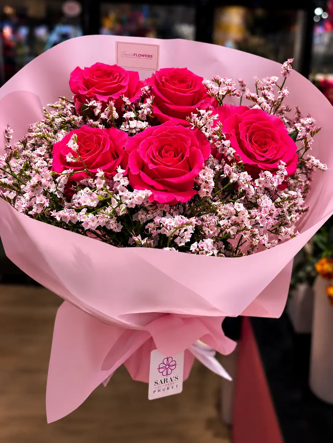A bouquet of 5 bright pink roses wrapped in light pink paper with a matching bow and tag, photographed in a real indoor setting with blurred background lights.