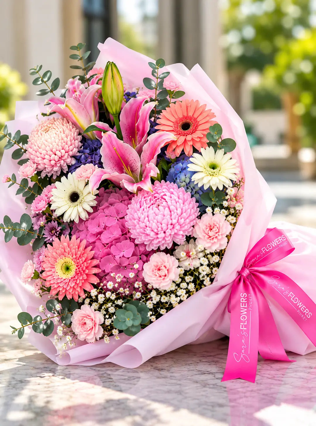 A vibrant mixed flower bouquet wrapped in light pink paper with a bright pink ribbon. The bouquet features pink and white lilies, pink and coral gerbera daisies, pink and white carnations, purple flowers, and green eucalyptus leaves, set against a softly blurred outdoor background.