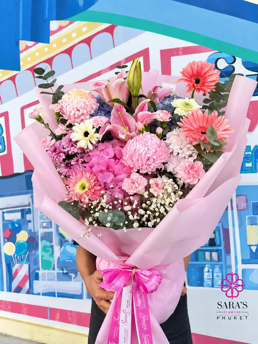 A vibrant, multicolor flower bouquet wrapped in pink tissue paper and tied with a pink ribbon, held by a person in front of a colorful, cartoon-style storefront backdrop. The bouquet features pink carnations, pink gerbera daisies, orange gerbera daisies, purple hydrangeas, white baby's breath, and green eucalyptus.