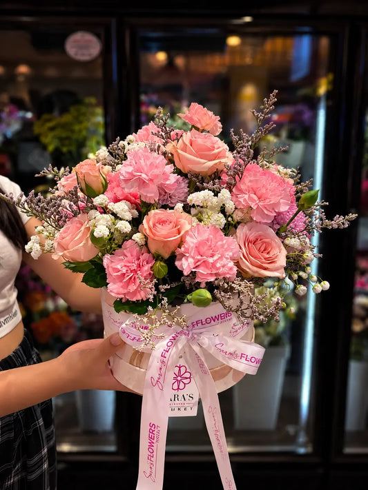 A large, vibrant floral bouquet held by a person in a flower shop. The bouquet features pink roses, pink carnations, white baby's breath, and green foliage, tied with a pink ribbon printed with the brand name 'JAV'S FLORIST'.