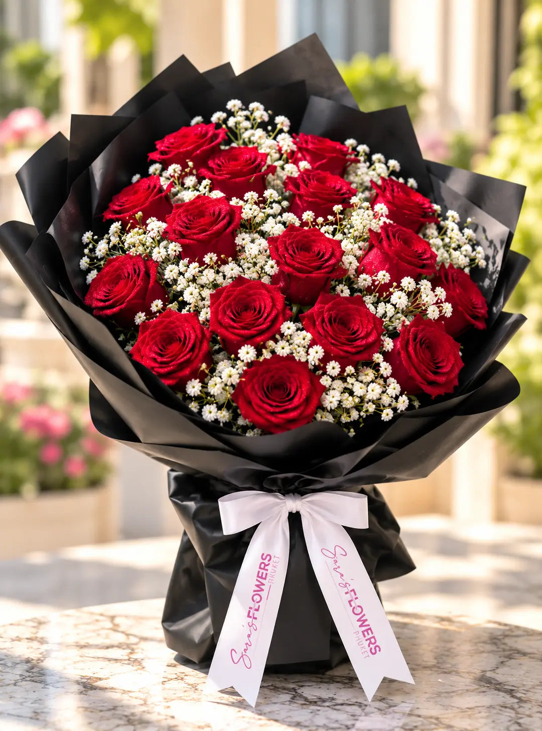 A bouquet of 21 red roses mixed with white baby's breath, wrapped in black paper with a white ribbon printed with 'Sara's Flowers' on the ends, set on a marble surface with a blurred garden background.
