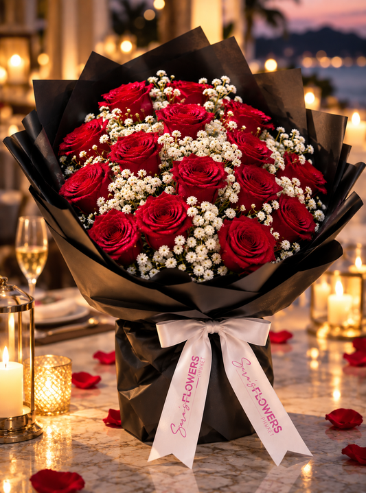 A large bouquet of red roses and white baby's breath wrapped in black paper with a white ribbon printed with 'Super Flowers', displayed on a candlelit table with rose petals and blurred background lights.