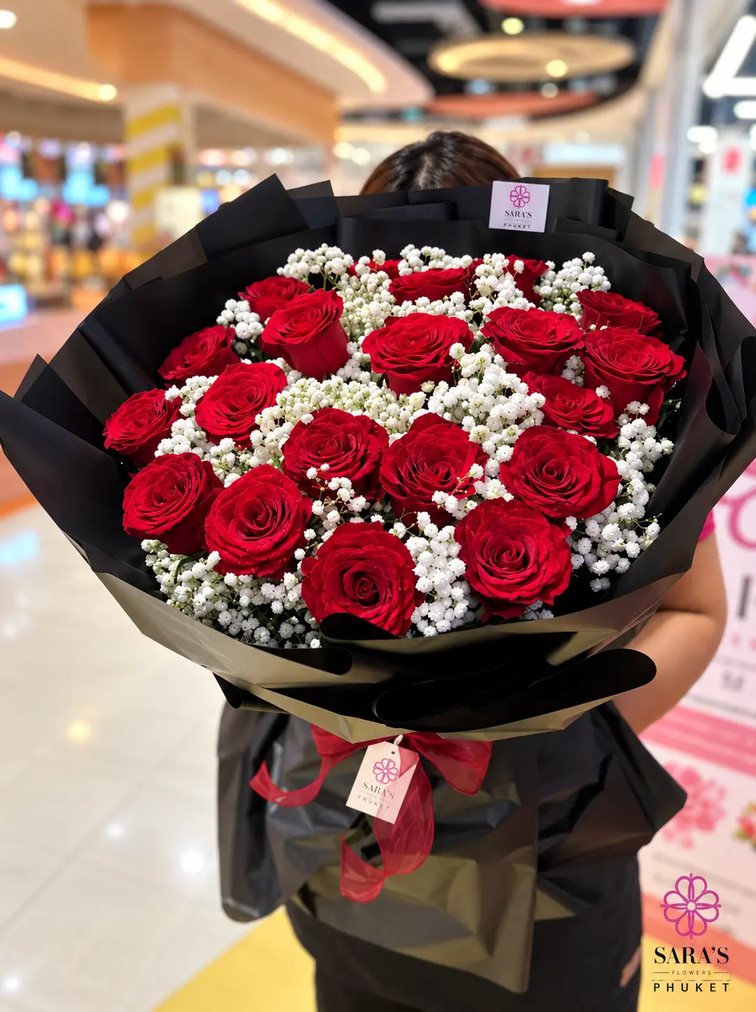 A large bouquet of red roses and white baby's breath flowers wrapped in black paper with a red ribbon, held by a person in a brightly lit indoor shopping mall setting.