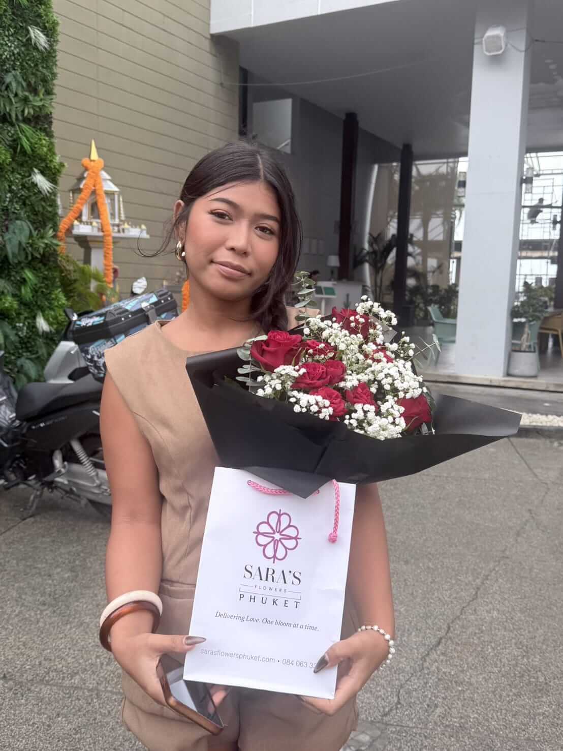 Woman holding a bouquet of flowers and a card with 'Sara's Flowers' branding outside a building.