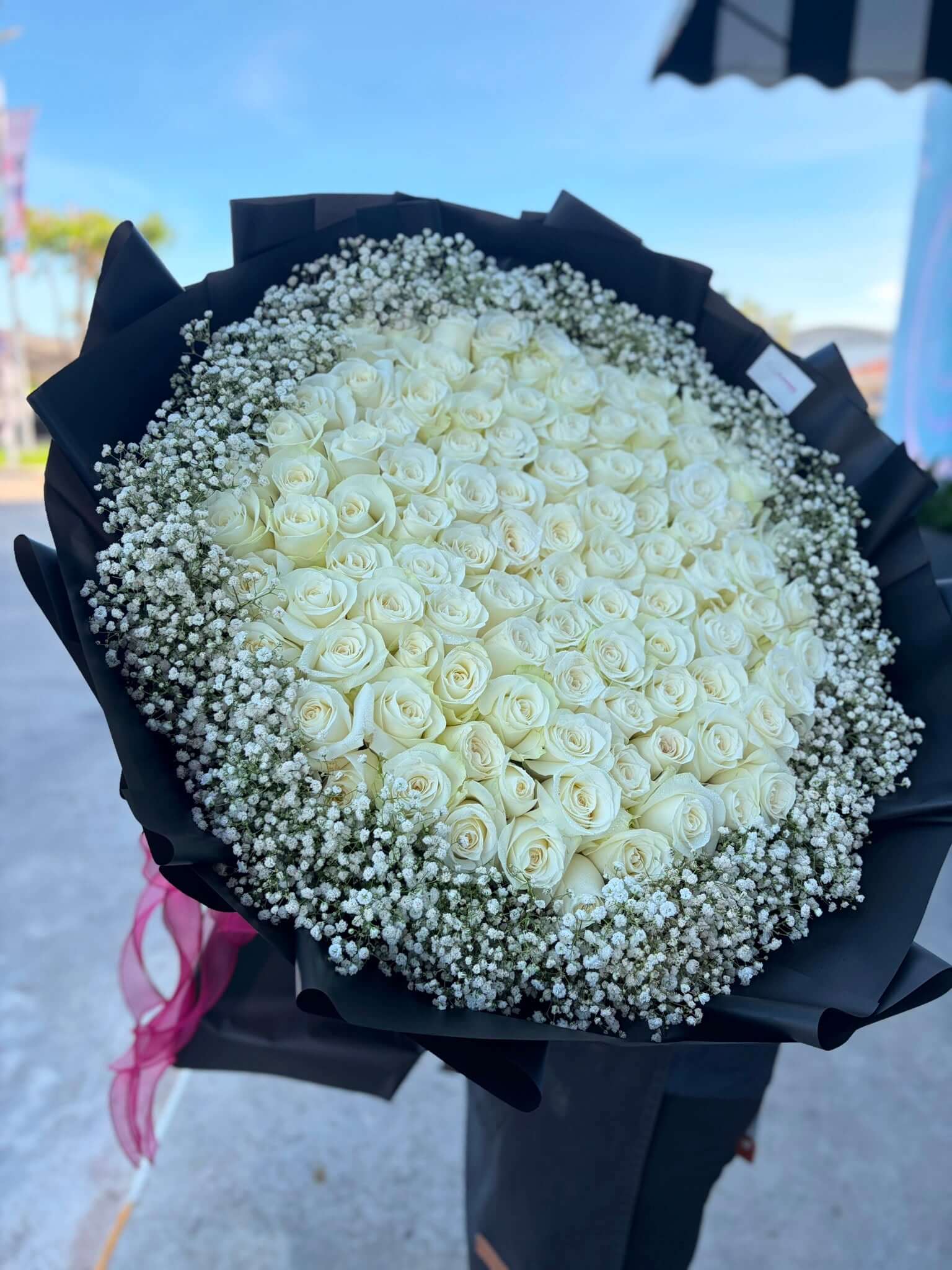 Bouquet of white roses with baby's breath in a black wrap, held outdoors.