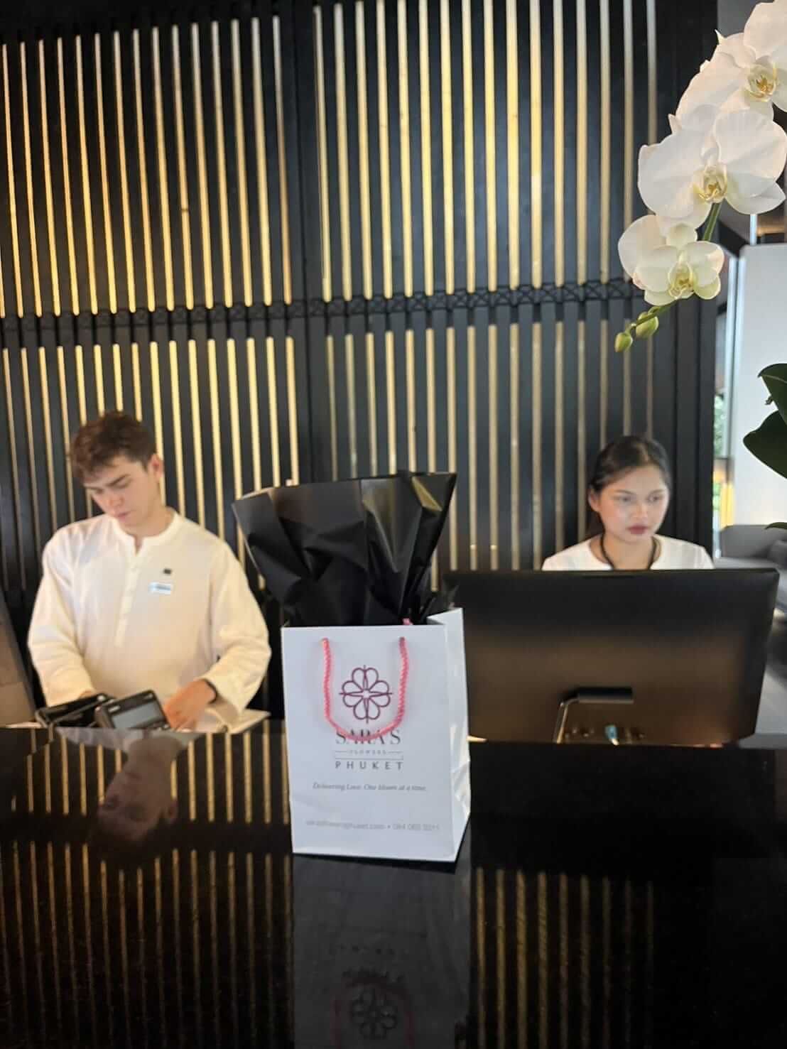 Two people working at a reception desk with a flower gift bag and black bag on the counter.