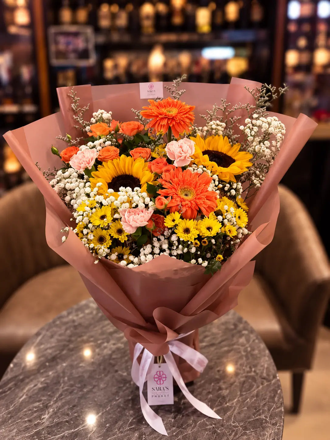 A vibrant flower bouquet wrapped in dusty pink paper, featuring sunflowers, orange and yellow gerbera daisies, pink roses, and white baby's breath, tied with a white ribbon and a 'MELVIN FLOWERS' tag, set on a marble table in a warmly lit shop.