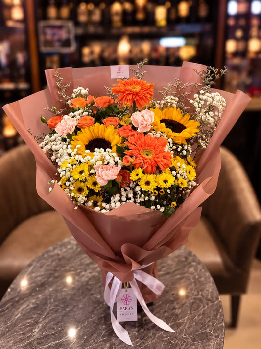 A vibrant flower bouquet wrapped in dusty pink paper, featuring sunflowers, orange and yellow gerbera daisies, pink roses, and white baby's breath, tied with a white ribbon and a 'MELVIN FLOWERS' tag, set on a marble table in a warmly lit shop.